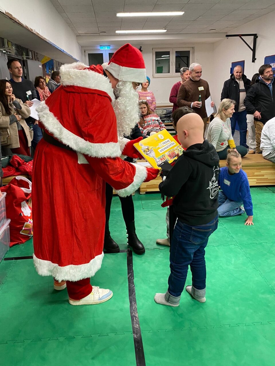 Ein Weihnachtsmann übergibt einem Kind ein Geschenk. Der Raum ist eine Turnhalle. Die anderen Kinder und ihre Eltern stehen dahinter und schauen zu.